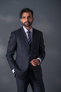 Man in a navy pinstripe suit and tie standing with hands in his pockets against a dark, moody background.Model in an embellished strapless gown with feather detailing, one arm raised, against a pale studio backdrop.