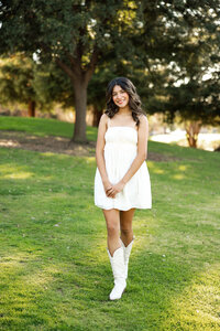High school senior girl standing in a grassy park wearing a white dress and boots, natural light senior portrait by Kaitlyn Dawn Photography.