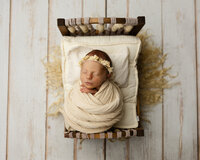 Peacefully sleeping newborn baby wrapped in a neutral cream swaddle and wearing a floral headband while posed on a wooden baby bed during a newborn photography session by Kaitlyn Dawn Photography.