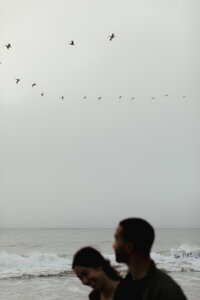 A blurry couple is seen in front of a cloudy shore as birds fly overhead. Photo taken at Baker Beach in San Francisco by Jordan Katz.