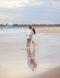 In Vilano Beach, a couple stands close in the shallow surf, foreheads nearly touching, their reflection mirrored beneath them while a sailboat glides across the horizon behind. - A portrait experience by Nature's Child Photography.