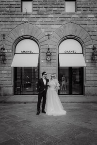 Mariés posant élégamment devant une façade historique aux arches jumelles, robe vaporeuse et complet noir, ambiance urbaine italienne en noir et blanc