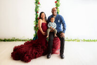 A man and pregnant woman in a red dress happily pose with their adorable baby.