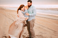 A pregnant mother smiles with her son as they press their foreheads together. Her husband smiles at them while holding he son from behind. The three are on a golden beach shortly after sunrise. The sky and water are pale blue with subtle hues of pink.