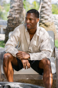 Cream linen shirt and dark shorts, seated in dappled light