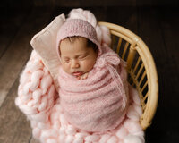 Newborn baby girl sleeping while wrapped in a soft pink swaddle and wearing a knitted bonnet while posed in a wicker basket with a fluffy pink blanket during a studio newborn photoshoot by Kaitlyn Dawn Photography.