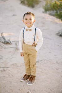 Young boy from the Collins family smiling in the Las Vegas desert, wearing suspenders and a bow tie during a golden hour children’s portrait session.