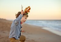 Young boy wearing reindeer antlers and a blue button-down shirt mid-jump near the shoreline during a beach child portrait session. (Photographed by Nature's Child Photography)