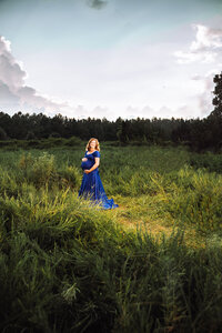 A redheaded pregnant woman in a royal blue dress stands in a large overgrown field on a cloudy day.