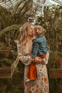 Mom wearing a floral dress holds little girl in rust bellbottoms and jean jacket while giving her a kiss in a Greenhouse family photoshoot