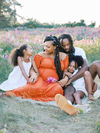 Pregnant mom, dad and two sisters sitting on a blanket in a meadow
