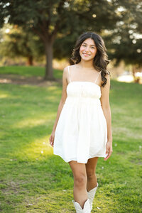 High school senior girl walking through a park wearing a white dress and boots, outdoor senior photography by Kaitlyn Dawn Photography.