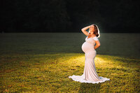 A backlit pregnant woman stands in a short cut grassy field smiling up at fireflies.