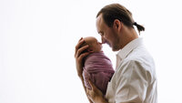 A backlit picture of a father nose to nose with his newborn daughter. The father wears a white shirt and the daughter is wrapped in a purple layer.