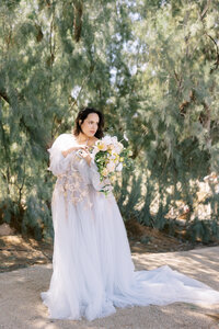 Pregnant woman in a flowing, ethereal gown holds a bouquet of soft pastel flowers during an outdoor maternity portrait session, surrounded by lush greenery in a light and airy setting.