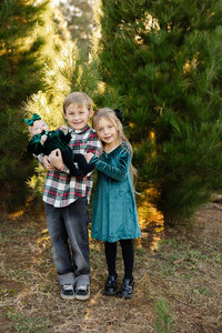 Two siblings standing together while holding their little sister outdoors surrounded by greenery and warm sunlight, candid family photography by Kaitlyn Dawn Photography, professional family portrait studio.