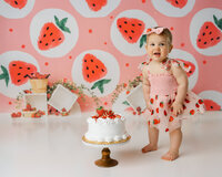 Baby standing beside cake in strawberry dress with watermelon-themed backdrop. Photograph by Yucaipa cake smash photographer Kaitlyn Dawn Photography.