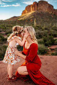 mother and daughter kneeling giving a kiss with hands on cheek with mountain backdrop