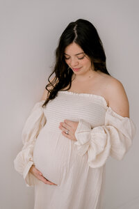 An expecting mom in a cream maternity dress, looking down at her belly while she holds it at Lake Arts Studio in Ohio.