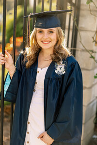 High school graduate wearing a blue cap and gown standing beside an iron gate in warm sunset light, senior portrait by Kaitlyn Dawn Photography.