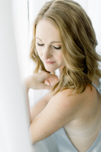 Close up photo of a woman posing by a window with sheer drapes with her hand elegantly places by her chest as she looks down over her shoulder for her boudoir session.