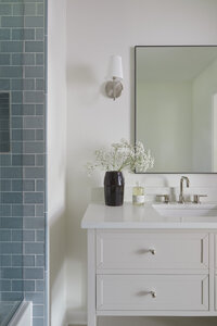 Bathroom with blue subway tile on the shower on the left. On the right is a single sink vanity in a neutral, beige paint with four drawers and an open shelf on the bottom. Above is mirror with a thin black frame and a silver sconces with a white shade on either side of the mirror.