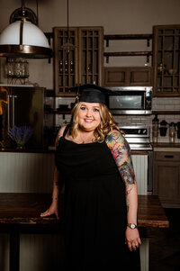 Kent State University online degree Nashville session at private home. Senior wearing black dress and cap in kitchen.