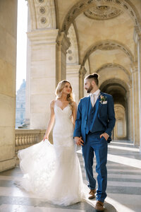 Bride in a lace gown and groom in a blue suit walking hand in hand beneath sunlit arches at a grand Paris venue.