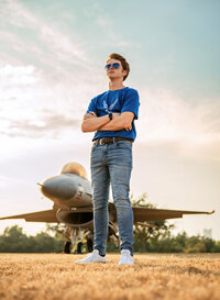 Senior portrait at Battleship Memorial Park in Mobile, Alabama— low angle with arms crossed with a sunset and an f-16 fighter jet in the background.