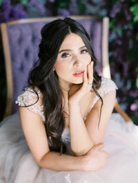 bride sitting on a velvet chair with a lilac floral installation behind her looking directly at the camera
