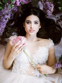 bride lying among peonies and lilac flowers holding a single bloom in a soft romantic editorial portrait