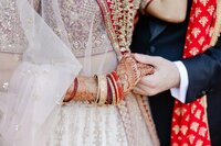 A close-up of a bride and groom in traditional south asian wedding attire, holding hands, showcasing intricate henna designs and jewelry.