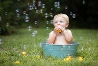 Jacksonville, FL portrait session capturing Blonde toddler sitting in a green metal tub on grass holding a small rubber duck as bubbles float around him during the splash portion of a cake smash in Mandarin.