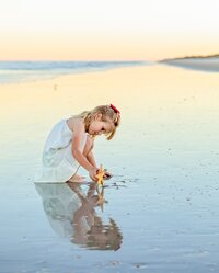Young girl in a white dress kneeling in shallow water and examining a starfish as her reflection appears in the wet sand during a child portrait session on the beach. (Photographed by Nature's Child Photography)