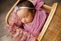 Sleeping newborn baby girl wrapped in a dusty rose swaddle and posed in a wicker chair prop during a studio newborn photography session by Kaitlyn Dawn Photography in Yucaipa.