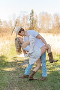 A country couple does a dip in a grassy field while sporting a cowboy hat and cowgirl boots for a styled shoot with a Duluth wedding photographer, S Jacobson Photography.