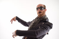 Performer in a dark jacket, goggles and patterned scarf with arms extended in a theatrical pose on white.