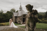 Mariés s'embrassant devant la chapelle de pierre du Château Ste-Agnès, statue en avant-plan flou, ciel nuageux et verdure du domaine viticole