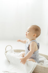 A baby boy sitting up in a basket on a studio floor while he looks and smiles at his mom.
