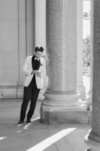 black and white image of groom fixing watch leaning against pillar outside in detroit