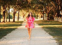 Senior portrait in downtown Magnolia Springs, Alabama — walking down a white gravel road wearing a bright pink outfit.