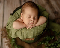 Sleeping newborn baby boy posed in a green bucket prop with hands tucked under chin and greenery accents during a studio newborn photography session by Yucaipa newborn photographer Kaitlyn Dawn Photography.