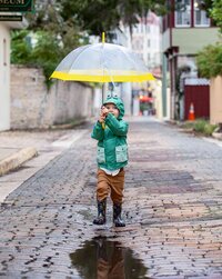 Toddler boy in a green raincoat and rain boots holding a clear umbrella while splashing through puddles on a wet cobblestone street during a child portrait session in the rain in St. Augustine. - A portrait experience by Nature's Child Photography.