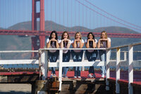 Five women dressed identically in blue jeans and white shirts leaning on a white railing with the golden gate bridge in the background