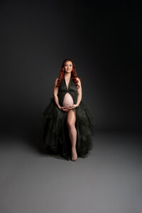 Expecting mother with long red hair wearing an elegant dark green maternity gown while smiling softly and cradling her baby bump during a fine art studio maternity photography session on a dark background by Kaitlyn Dawn Photography.