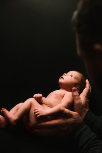 A naked newborn baby rests in her fathers hands looking up to an unseen light. She appears to float in his hands against a black backdrop.