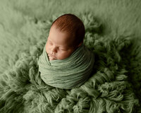 Sleeping newborn baby boy wrapped in a soft green swaddle and posed on a sage green backdrop during a studio newborn photography session by Yucaipa newborn photographer Kaitlyn Dawn Photography.