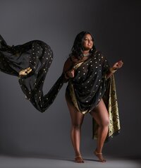 A woman draped in a flowing black and gold sari stands barefoot against a dark background. Her arm is raised, making the fabric billow dramatically—an image perfect for fashion-inspired editorial portraits Washington DC.