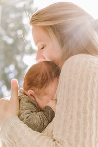 A baby snuggles into his mother's shoulder. They are backlit by a sunny, tree lined sky.