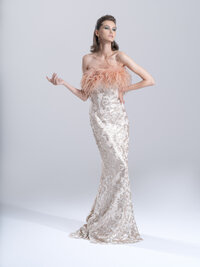 Model in an embellished strapless gown with feather detailing, one arm raised, against a pale studio backdrop.
